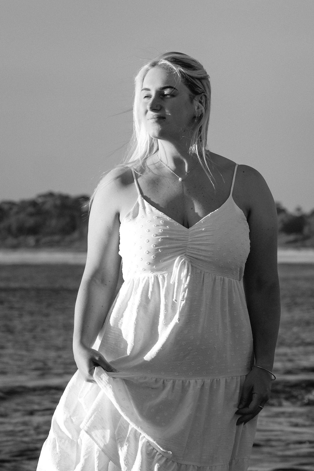 A series of black and white photos showing someone in a white sundress standing outdoors near water.