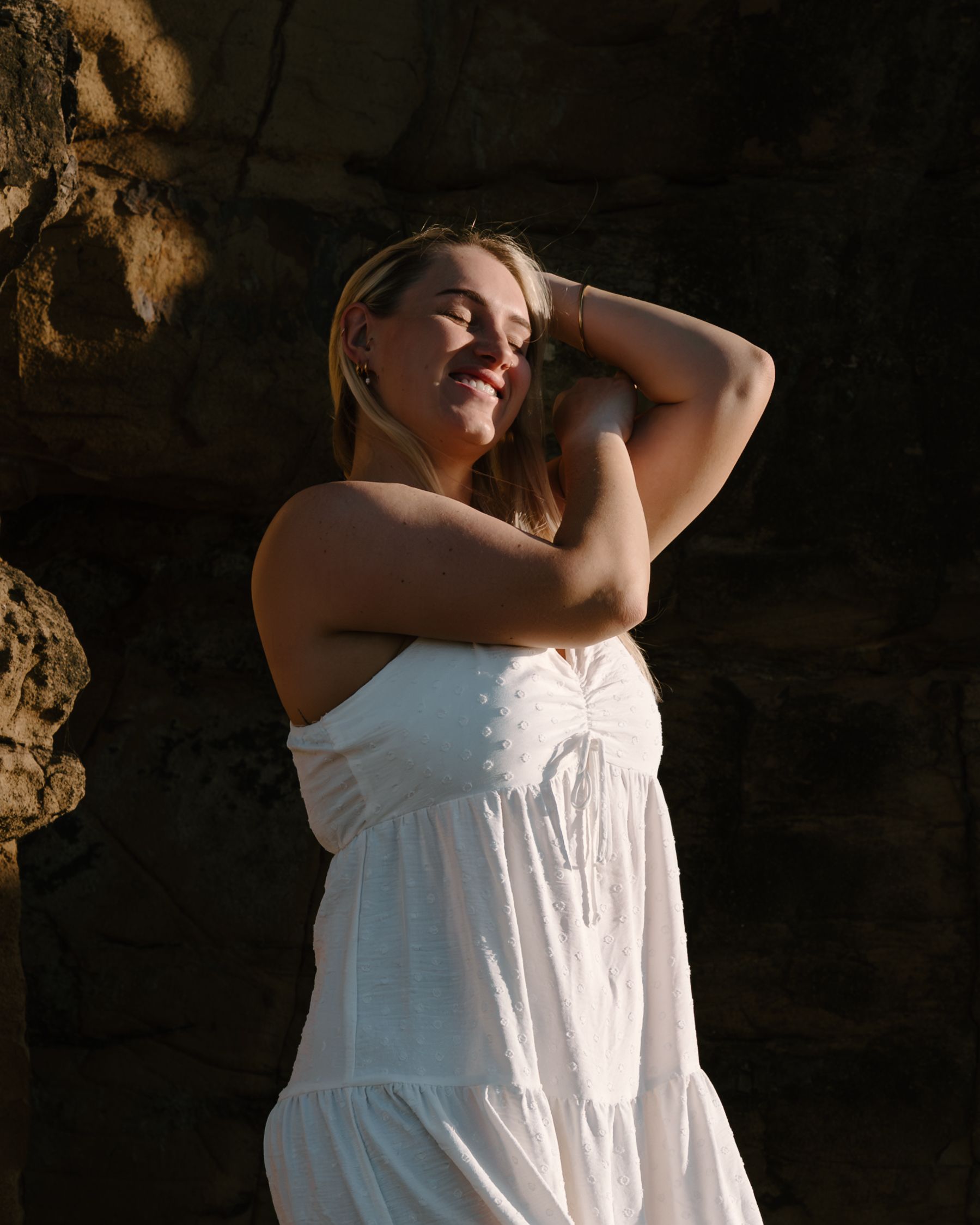 A person in a white summer dress poses against a rocky backdrop in warm sunlight.