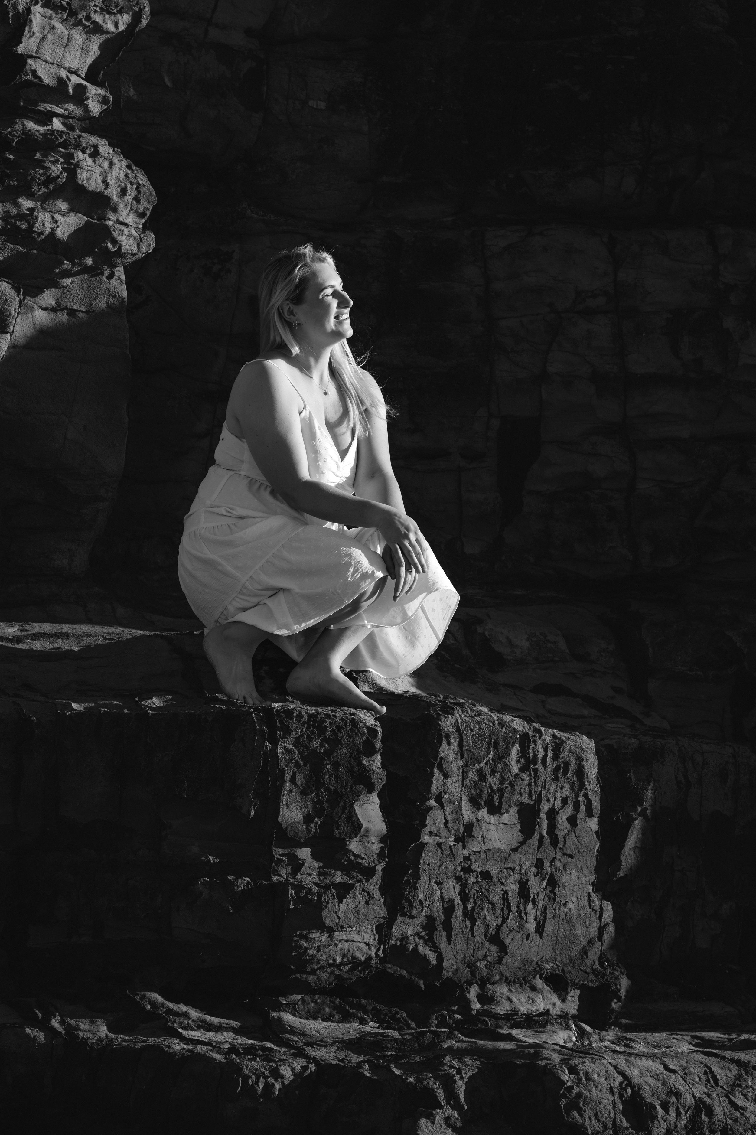 A figure in white clothing sits contemplatively on a rocky ledge in dramatic black and white lighting.