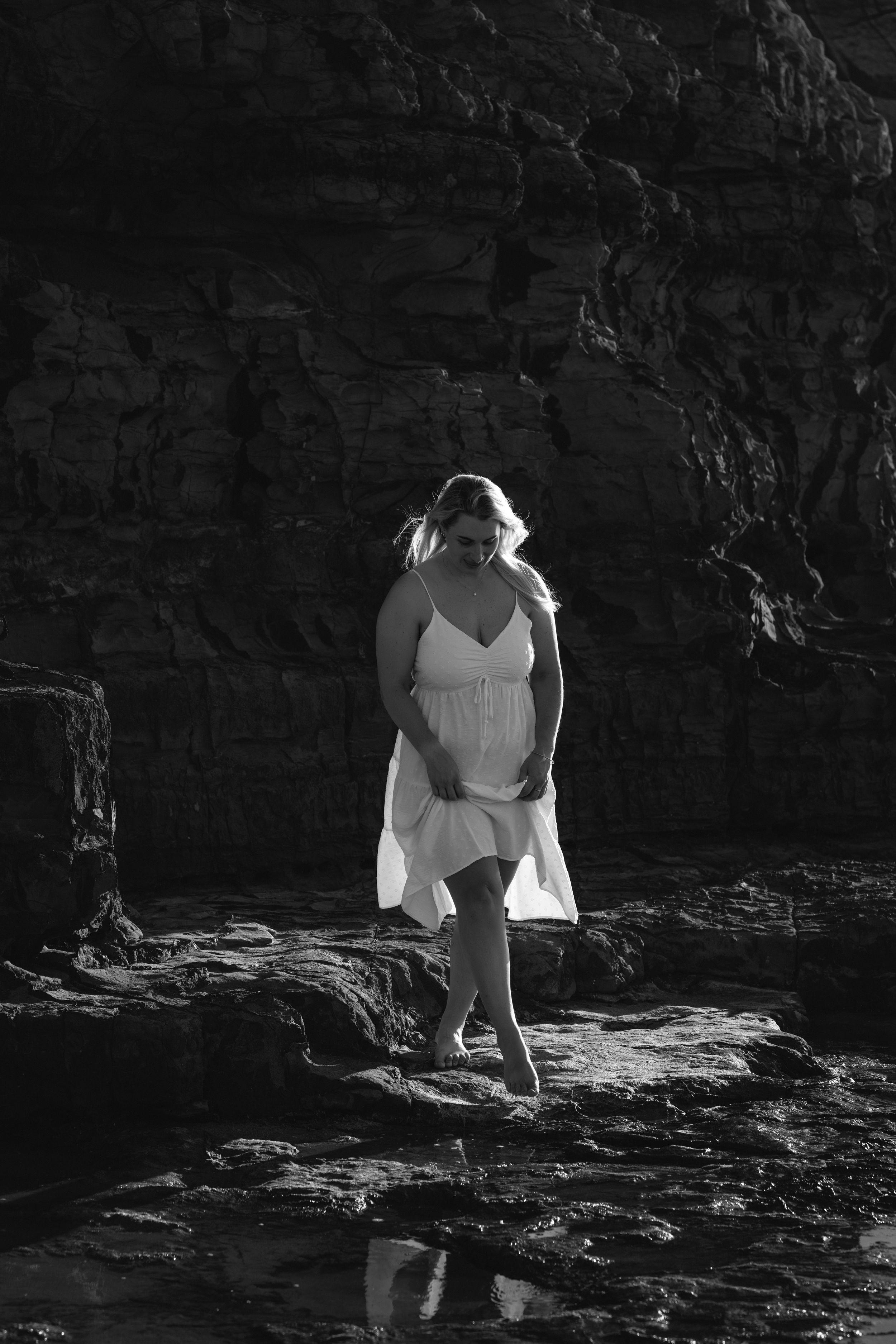 A person in a white dress crouches on dark rocky terrain in a dramatic black and white photograph series.