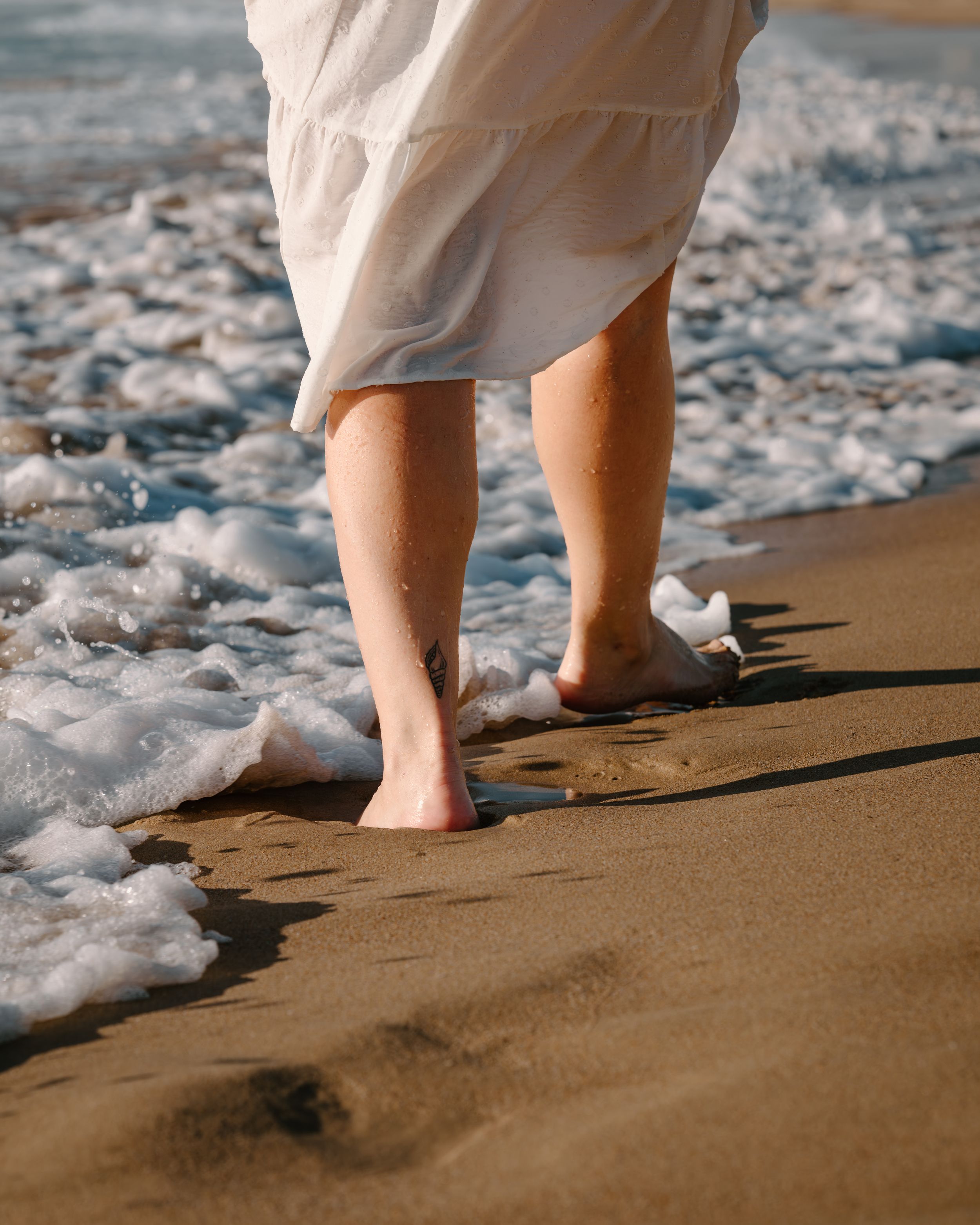 Bare feet walking on sandy beach with gentle ocean waves washing over the shoreline in warm sunlight.