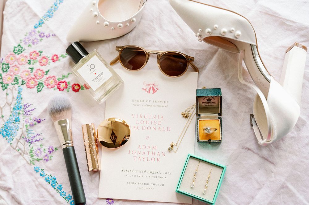 Wedding details flatlay with floral invitations, sunglasses and delicate accessories on white fabric.