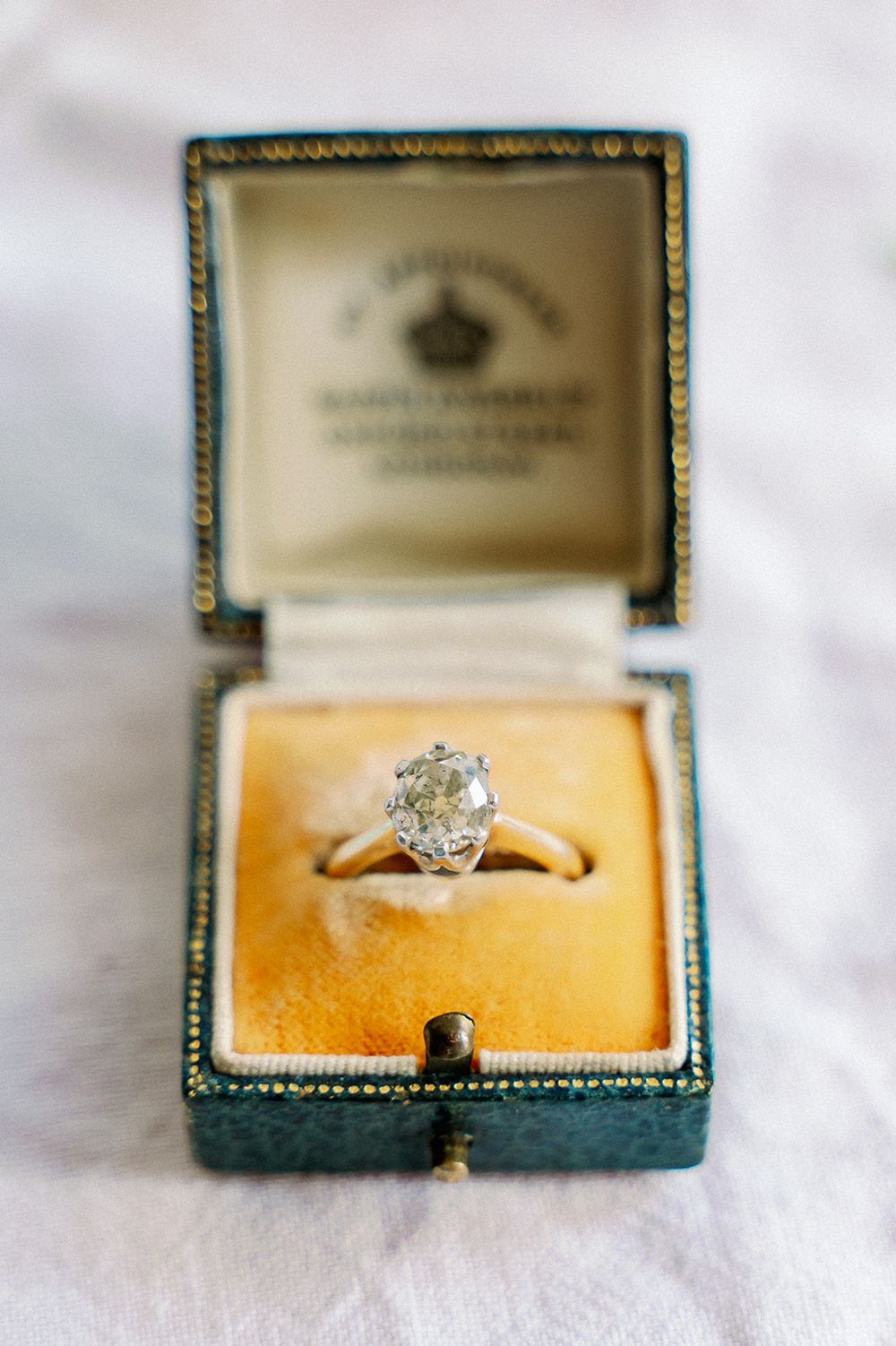 Vintage teal jewelry box displaying elegant diamond engagement ring on white fabric.