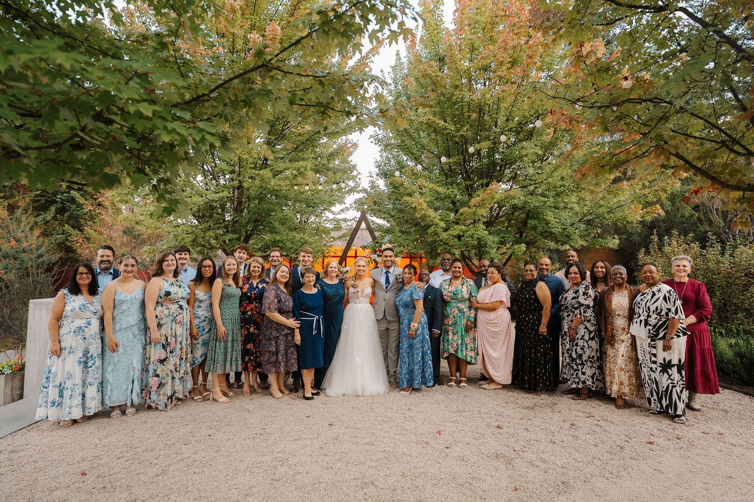 Wedding party guests line up for a panoramic group photo outdoors under tall green trees in autumn.