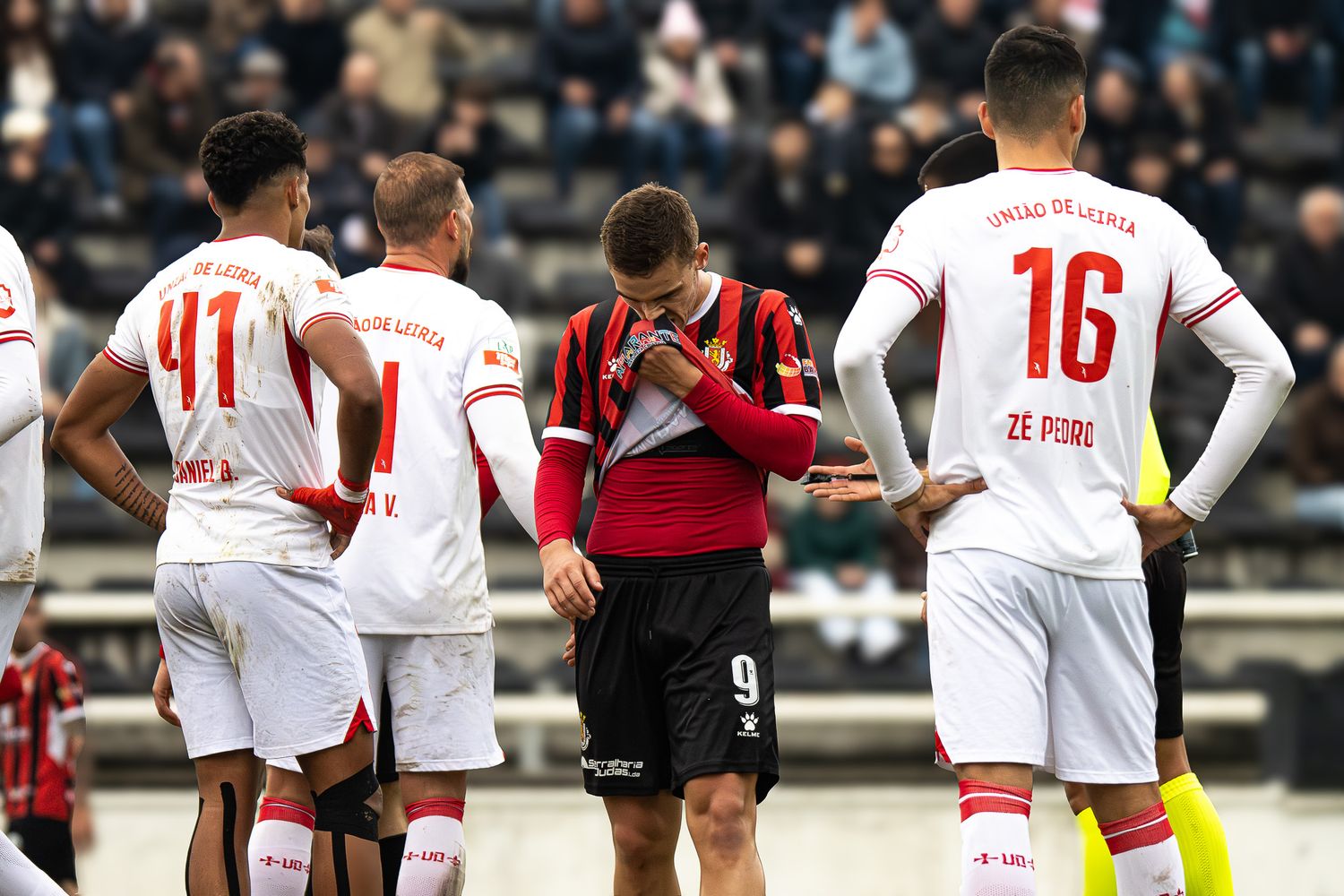 Soccer players line up on field during match with spectators visible in background.