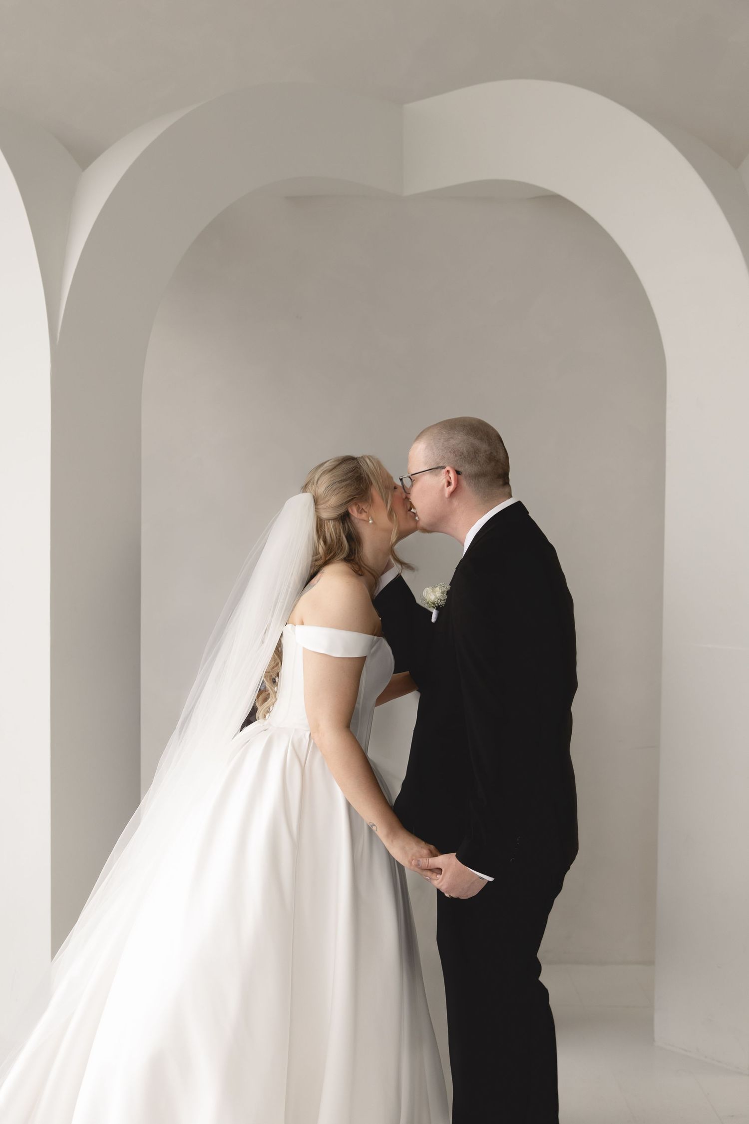 A romantic wedding couple embraces in a white arched hallway with minimalist architectural details.