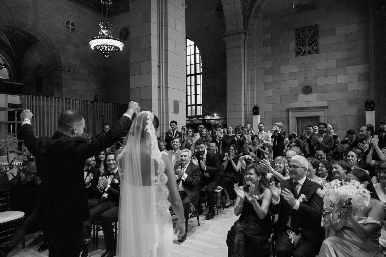 Black and white photo of a bride and groom celebrating their first walk as newlyweds, greeted by applauding guests in an elegant venue.