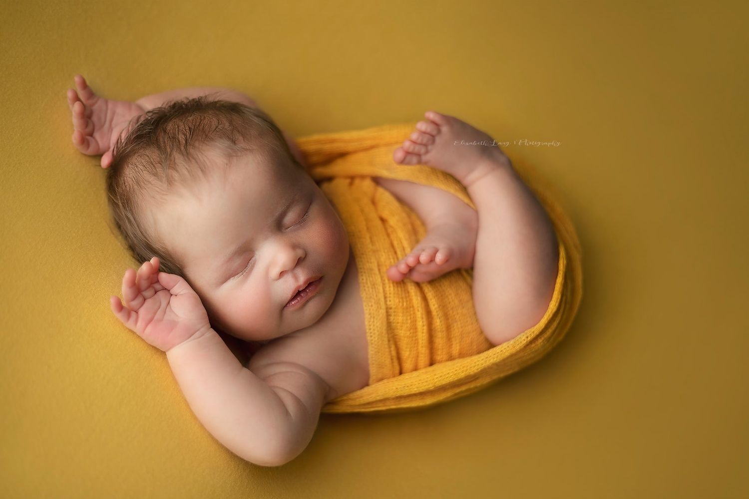 Newborn baby wearing yellow wrap sleeps peacefully against golden background in artistic portrait series.