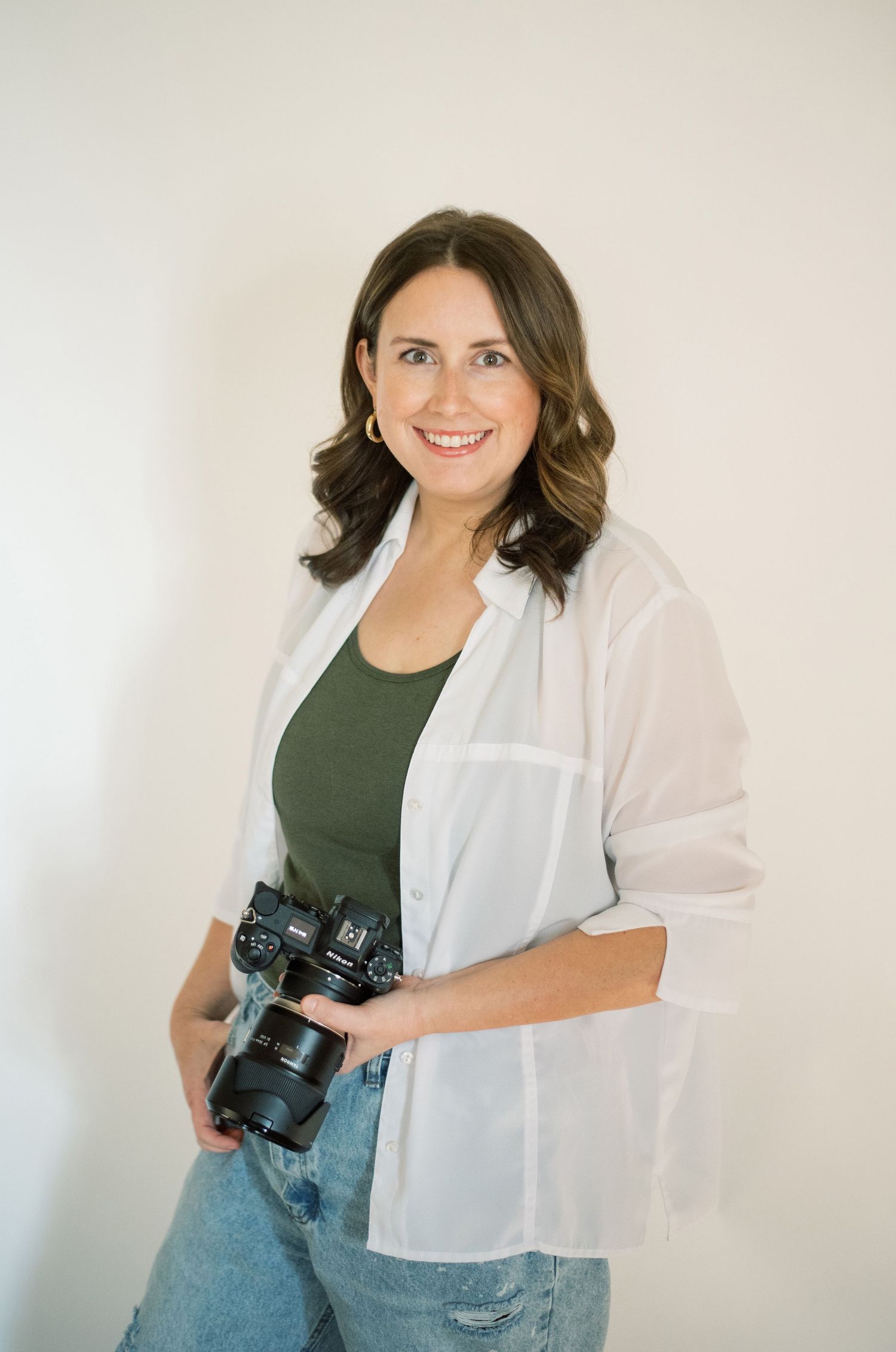 Professional photographer in white button-down and jeans poses with camera against white background.