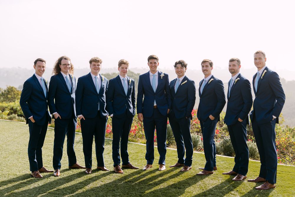 Wedding party in dark suits poses together on a grassy hillside during golden hour.