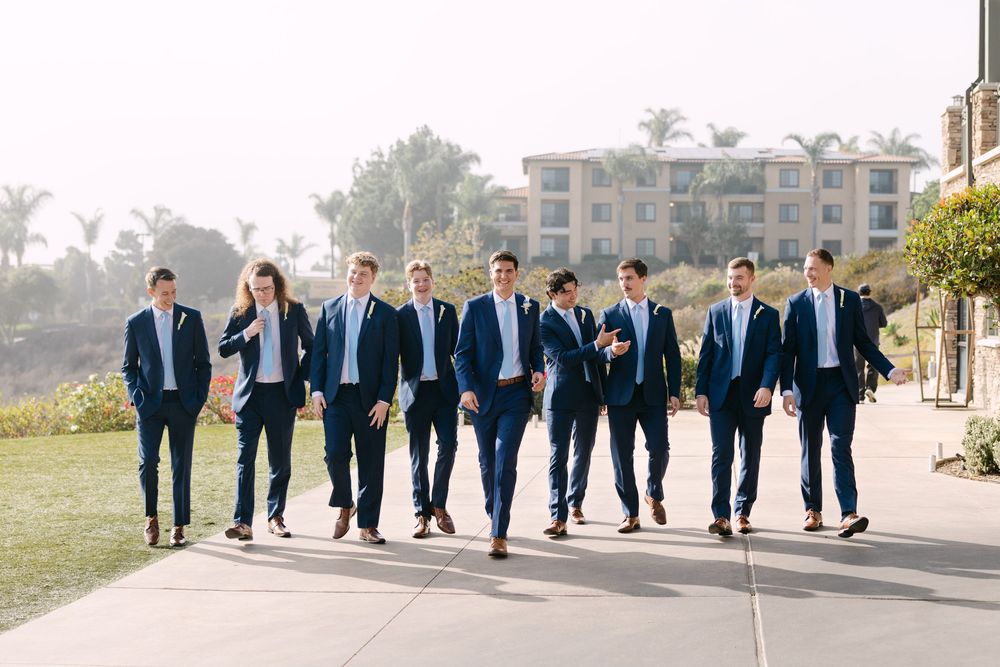 Wedding party members in navy suits and dresses pose in a line formation on a sunny day outdoors.