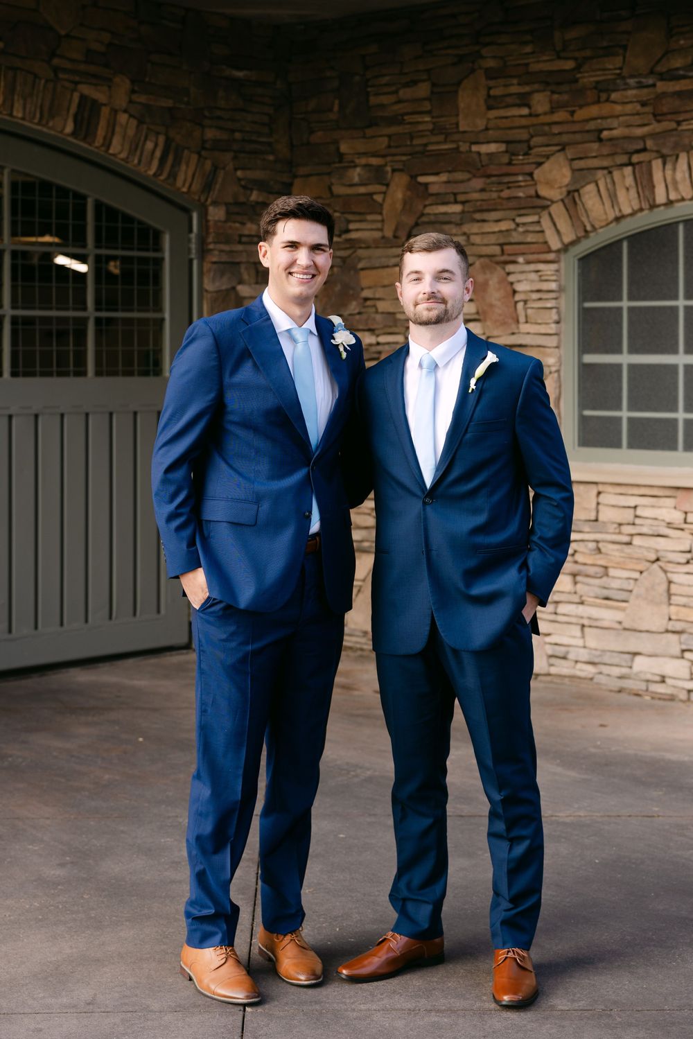 Groomsmen in matching navy blue suits and brown leather shoes stand in front of a stone wall and garage doors.
