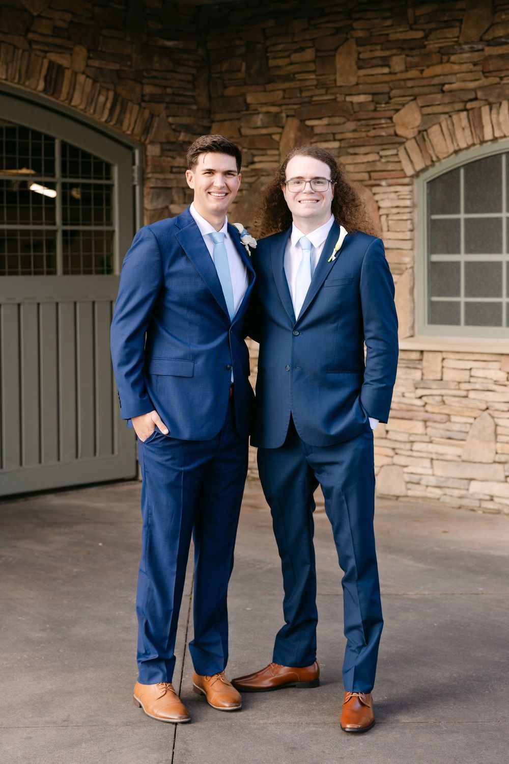 Two people in navy blue suits and brown shoes stand together in front of a stone archway entrance.