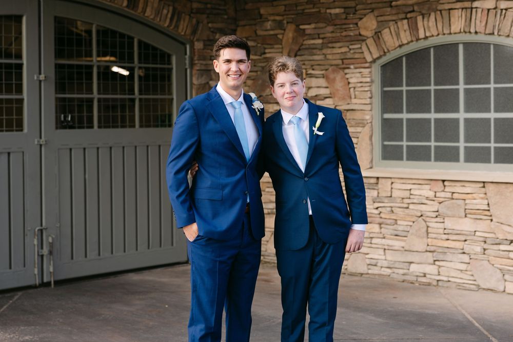 Two people in navy blue suits pose together in front of a stone wall and white garage doors.