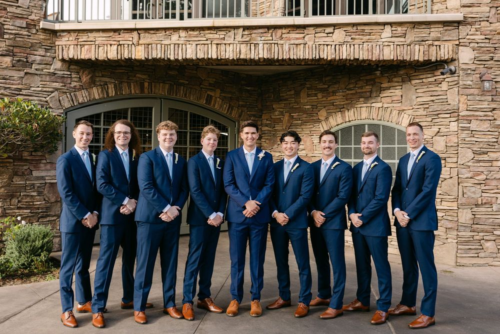 Groomsmen in navy suits pose in a row against stone wall backdrop at wedding venue.