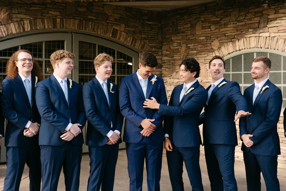 Wedding party in navy blue suits and light blue dresses pose together against a rustic stone wall backdrop.