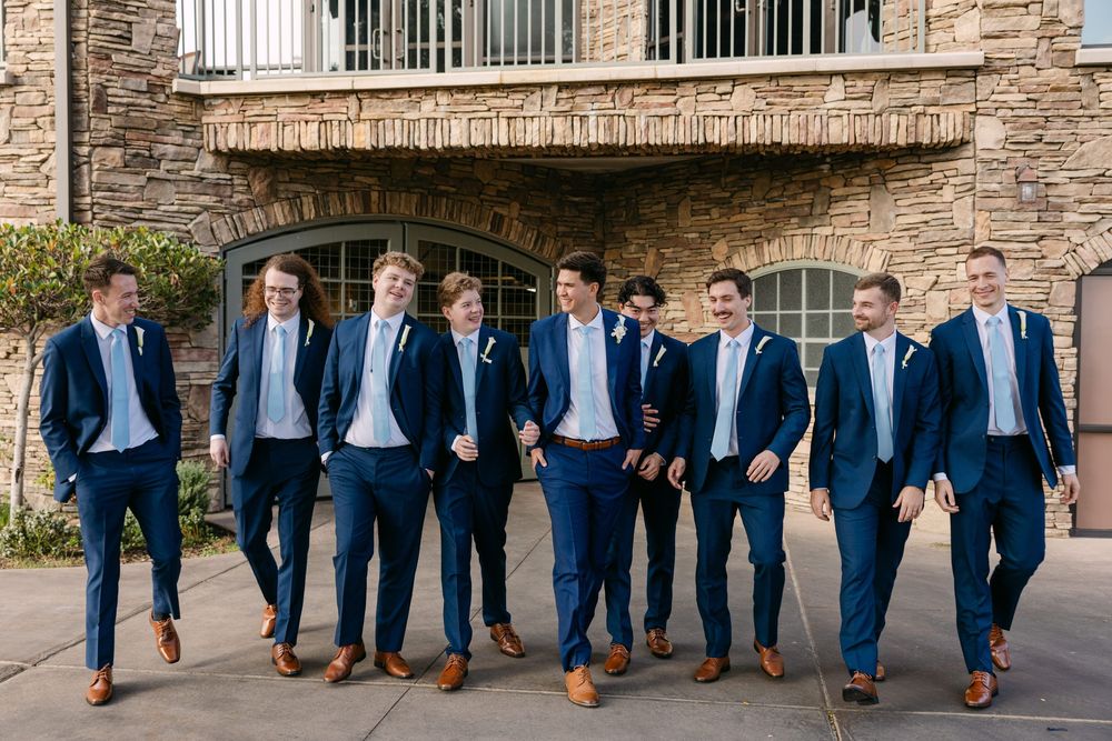 Groomsmen in navy suits and brown shoes walk together outside a brick building.