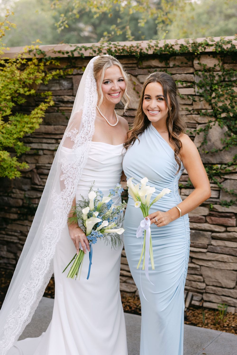 Wedding party members in light blue bridesmaid dress and white bridal gown pose together against stone wall outdoors.