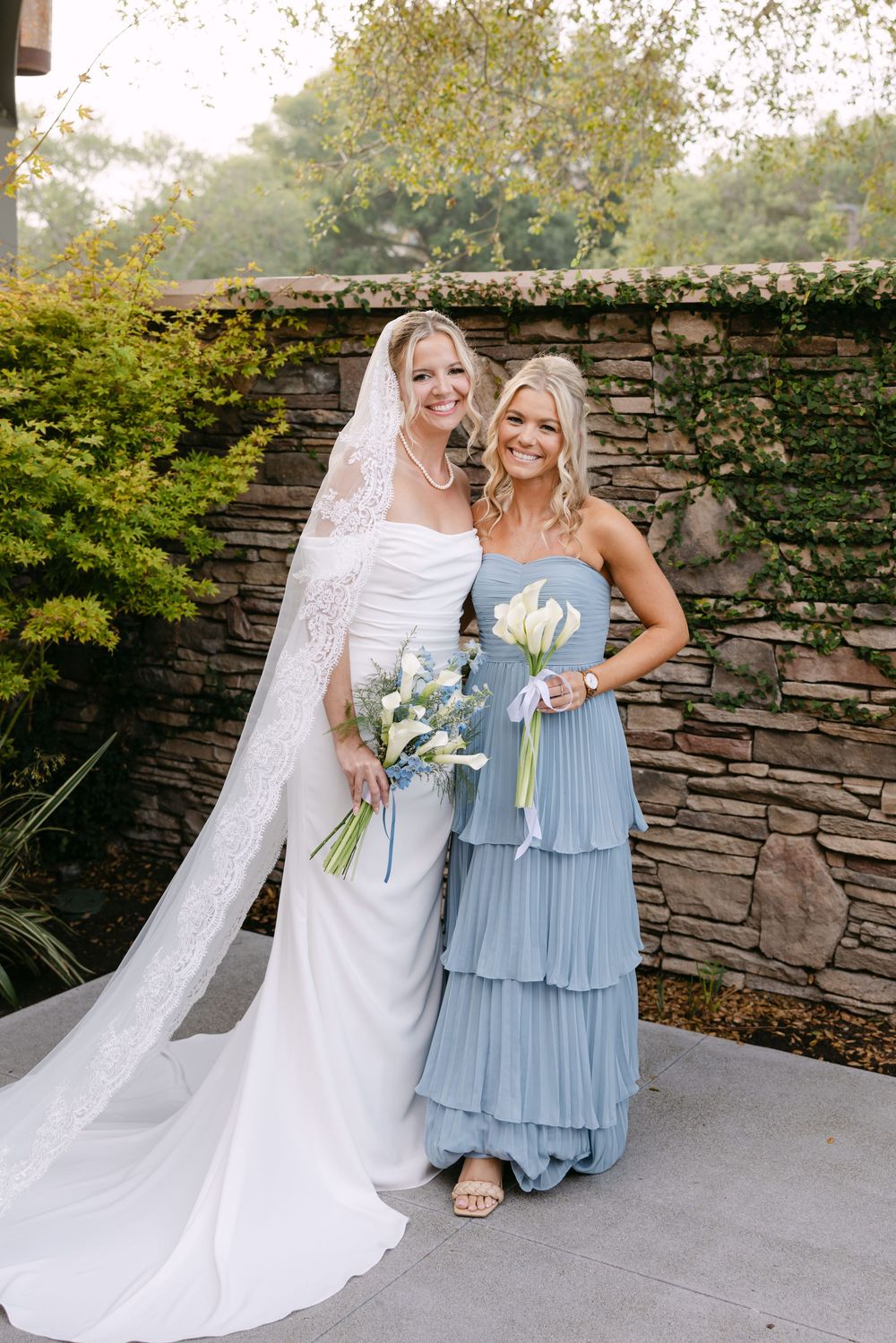 Bridal party members in light blue dresses and white gown stand by stone wall outdoors.