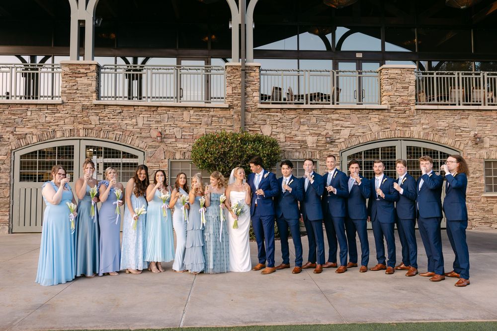 Wedding party in light blue dresses and navy suits poses together against stone building backdrop.