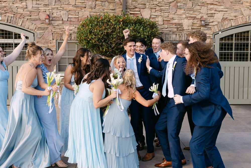 Wedding party members in blue attire dance and celebrate on stone patio.