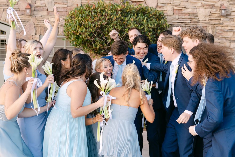 Joyful wedding party celebrates with dance moves in front of stone building.