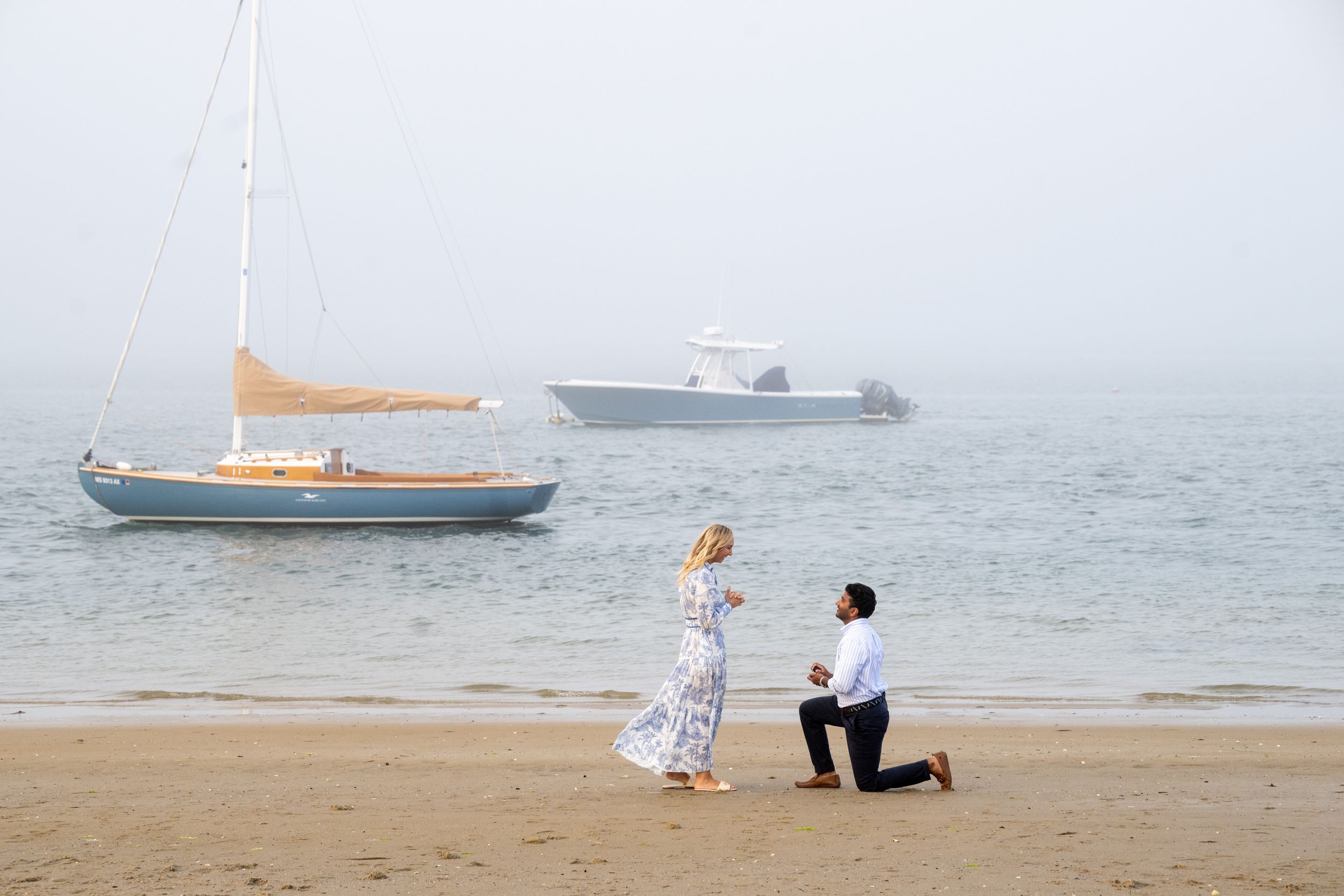 A scenic beach proposal with boats visible in the background across the misty water.