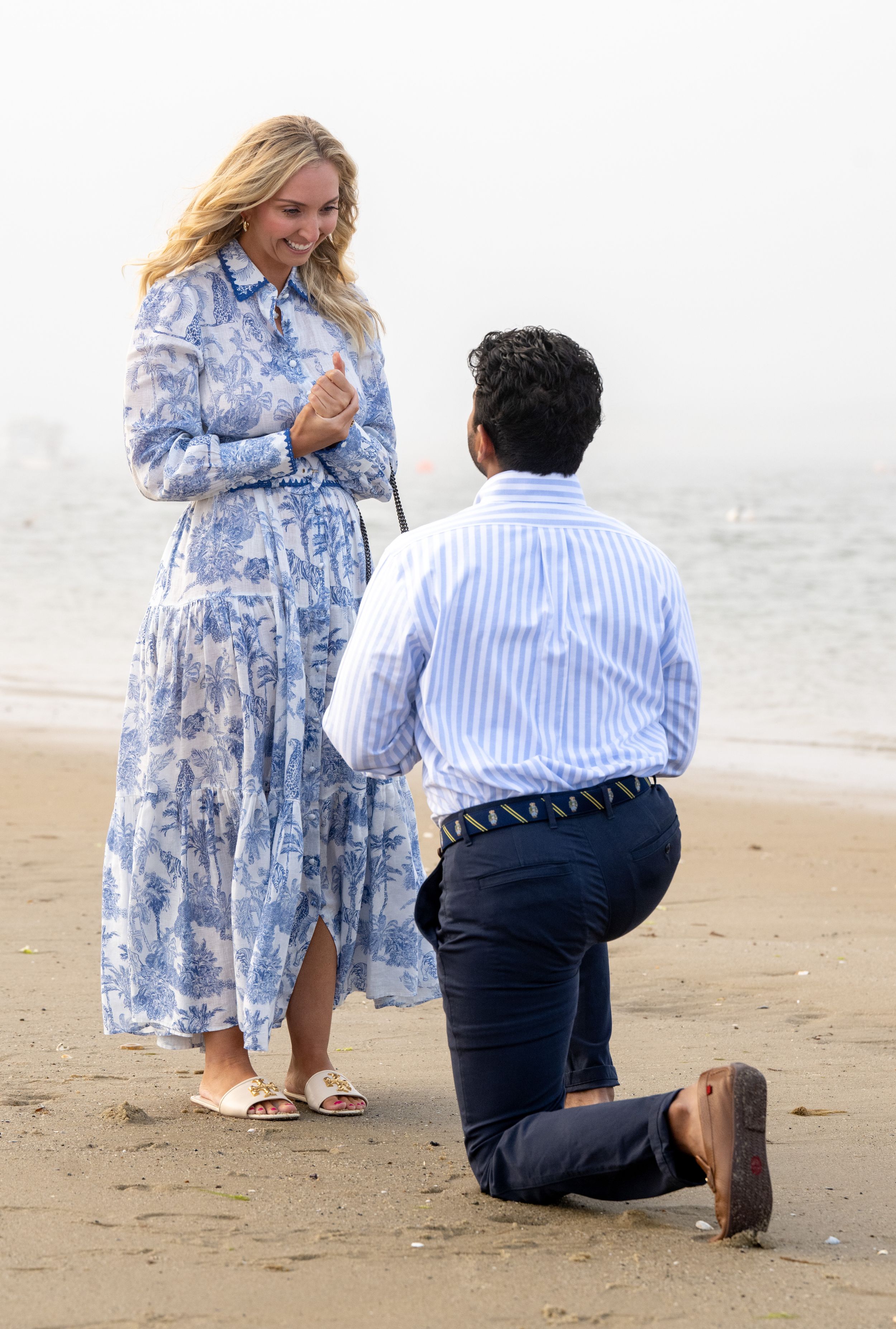A couple on the beach during a proposal moment with the woman in a floral blue dress.