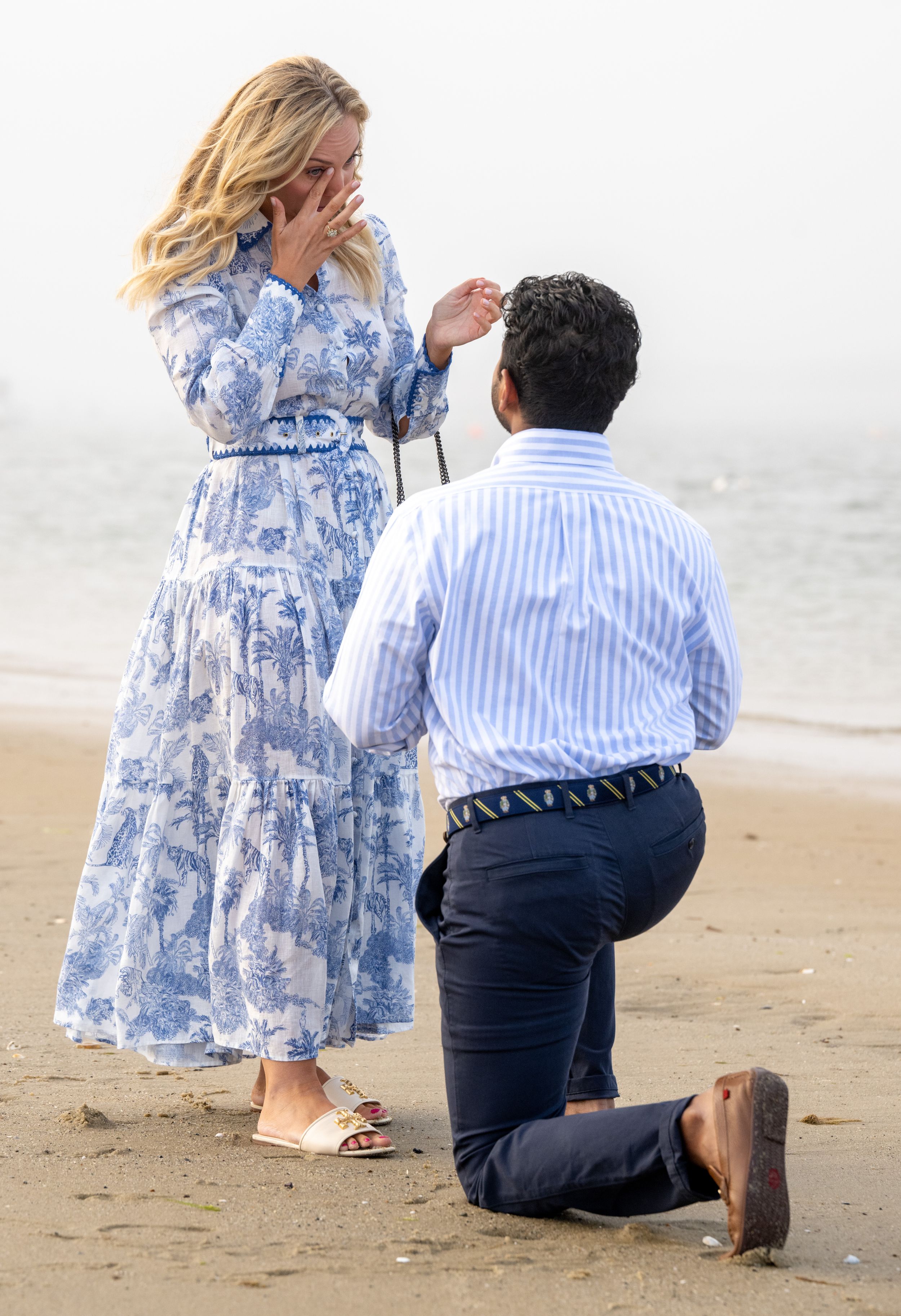 A proposal scene unfolds on a sandy beach with boats in the distance.