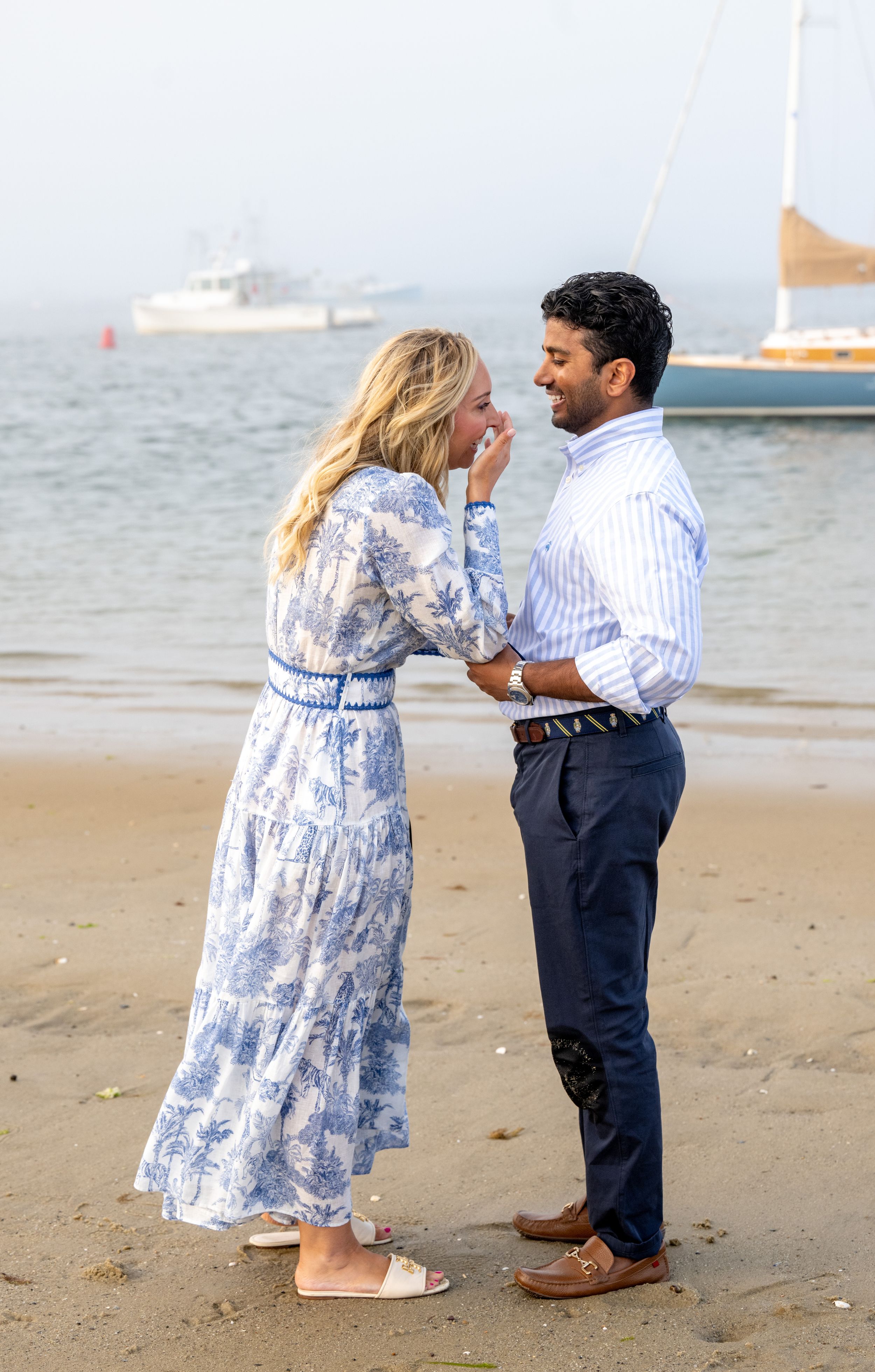 A couple celebrates their engagement on a beach with boats anchored in the distance.