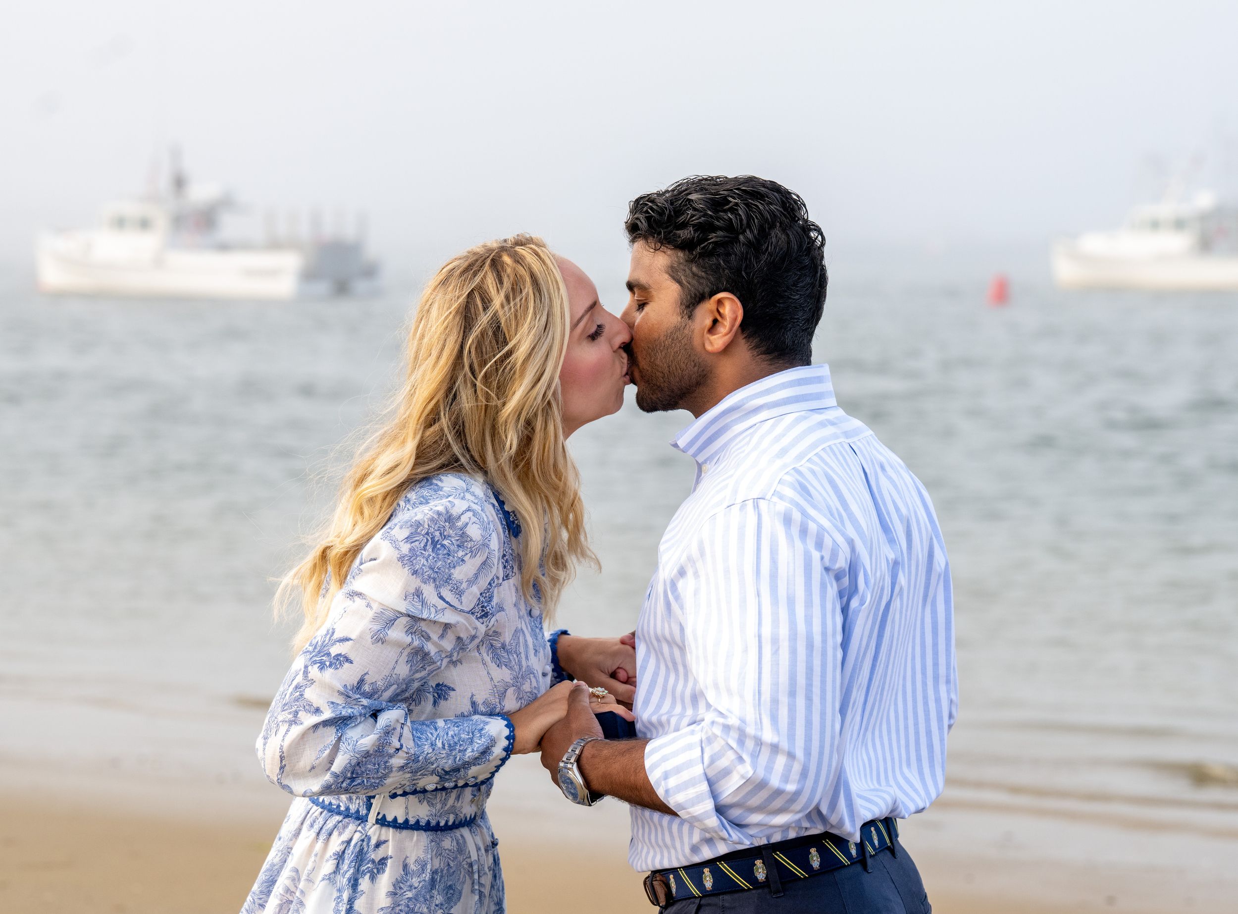 A romantic beach proposal scene with boats visible in the background waters.