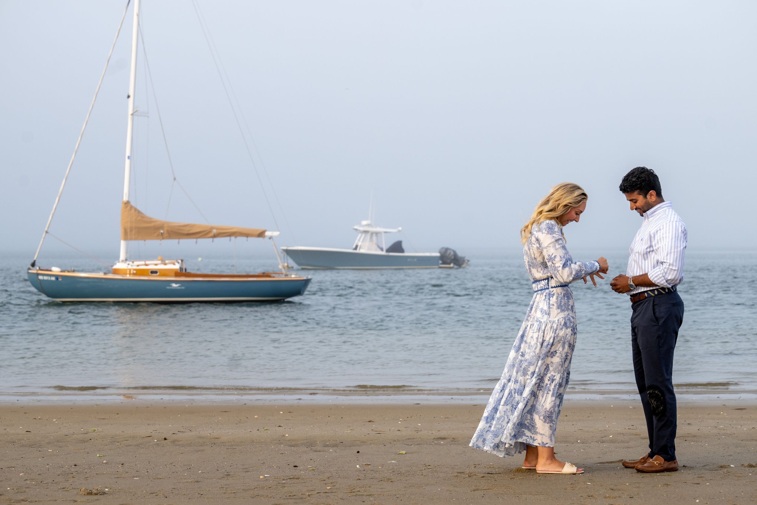 A couple shares an intimate moment on a beach with boats sailing in the harbor behind them.
