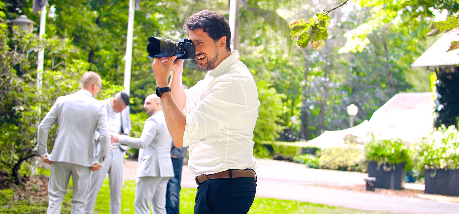 A man in a white shirt photographs outdoors at a garden event with guests in formal attire in the background.