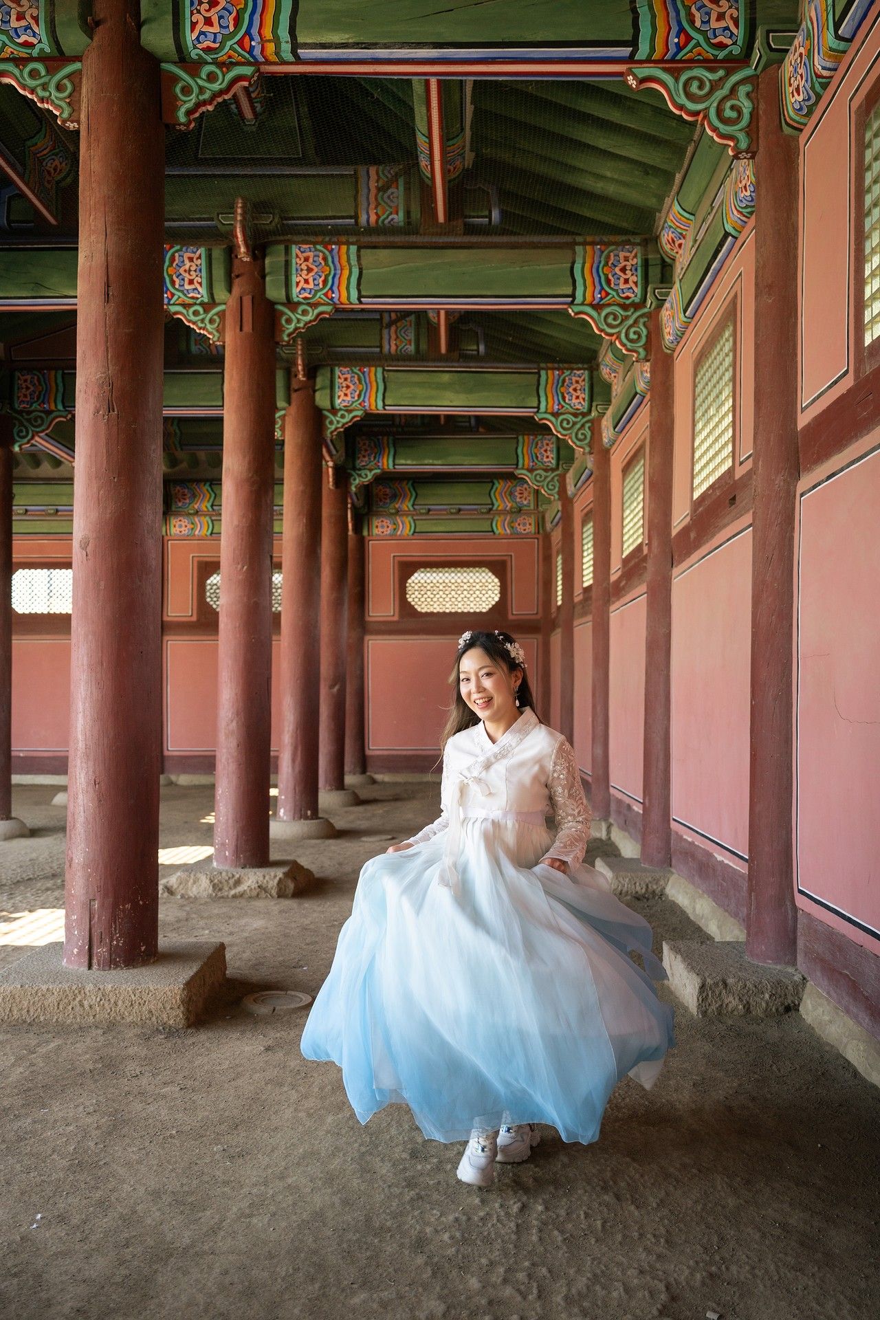 A person wearing a traditional Korean hanbok dress twirls gracefully under an ornate palace corridor with painted wooden beams.