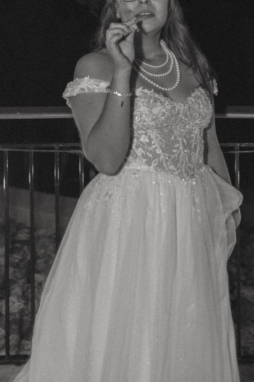 A bride in a white lace gown with pearl necklace poses against a dark evening backdrop.