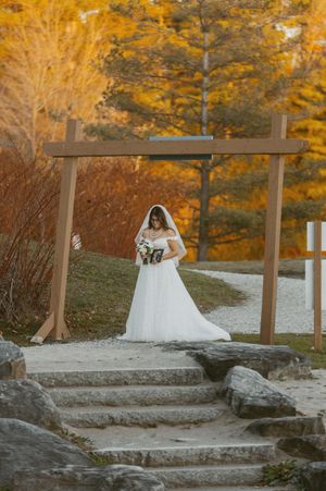 A bride in a white dress poses on stone steps with vibrant fall foliage in the background.