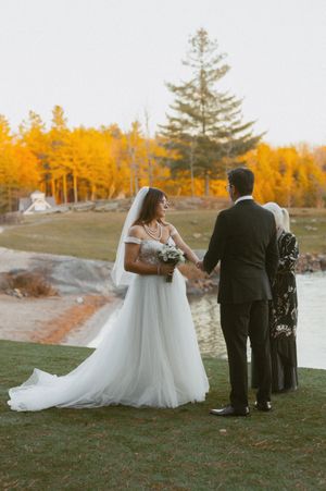 Wedding party gathers on a grassy hill overlooking a lake with golden autumn trees in the background.