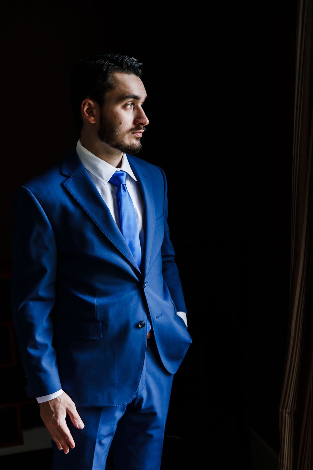 Groom in navy blue suit poses for dramatic portraits in natural window light.