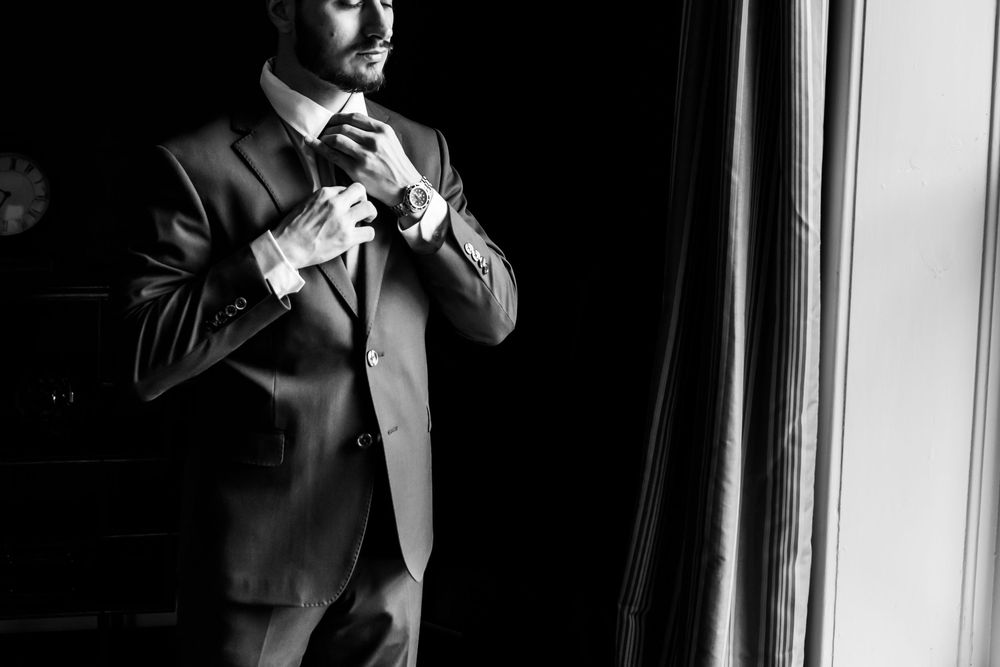 A person in a dark suit adjusts their tie while standing near a window in dramatic lighting.