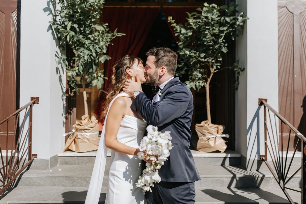 A couple shares a romantic moment in front of a white building with decorative greenery and floral arrangements.