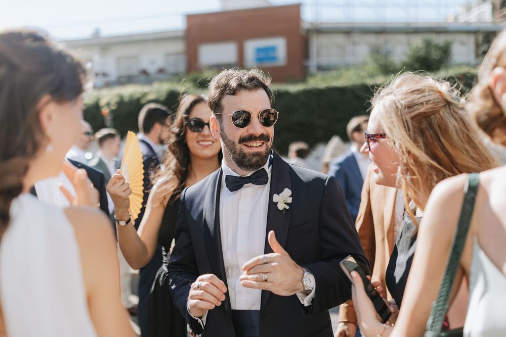 Wedding guests socialize outdoors during a sunny celebration while holding drinks and wearing formal attire.