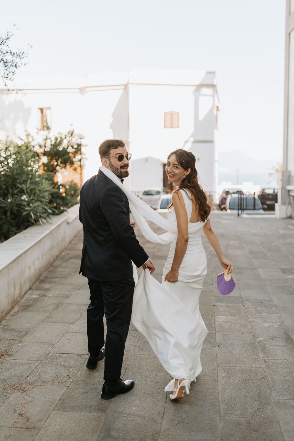 A couple in wedding attire dances playfully together on a stone terrace with ocean views at sunset.