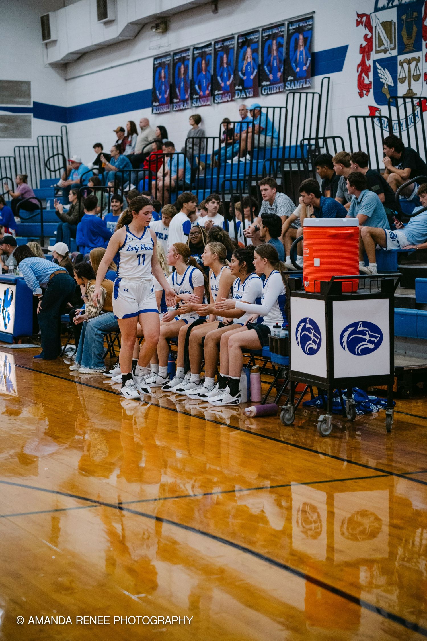 High school volleyball team sits on bench during match in blue and white gymnasium with spectators watching.