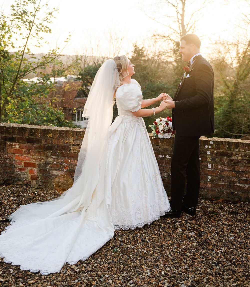 A romantic sunset wedding photoshoot featuring a couple in formal attire standing against a rustic stone wall.
