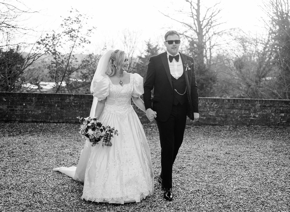 A black and white wedding photograph showing a couple walking together outdoors in formal attire.