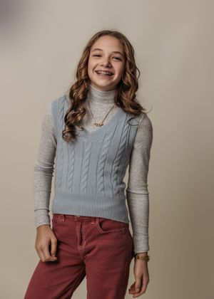 Child wearing blue sweater vest and burgundy pants poses against neutral backdrop.