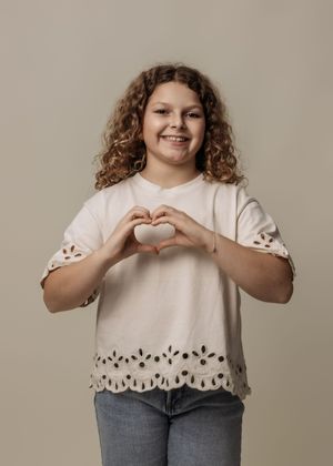 Child in white eyelet top makes heart shape gesture against neutral background.