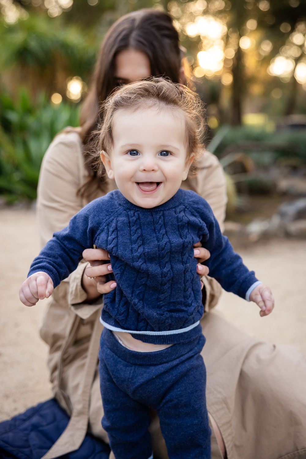 A baby smiles at the camera while his mom holds him from behind