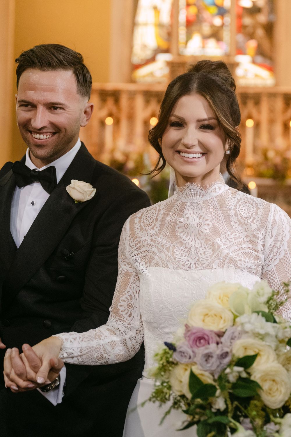 A bride in a lace high-neck wedding gown and groom in a black tuxedo smile during their church ceremony.