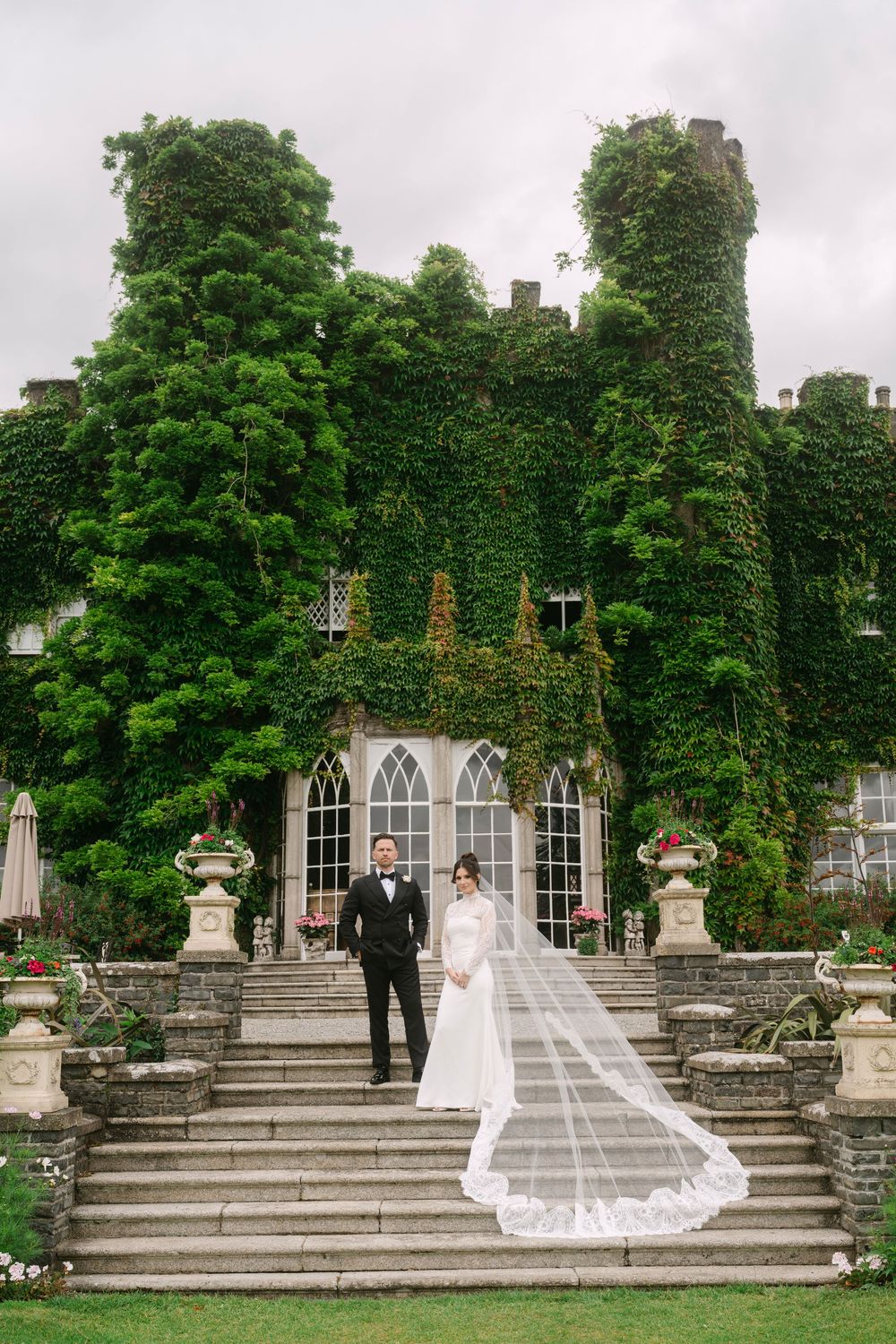 Wedding couple stands on grand stone steps of an ivy-covered historic building with Gothic architecture.